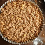 A freshly baked peach crumble in a ceramic dish with a golden, crispy topping, surrounded by fresh peaches and a serving spoon.